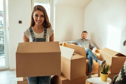 Woman in overalls holding a moving box while a man sits on the couch.