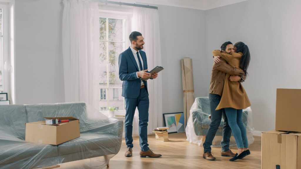 a young couple embraces in their new home as their real estate agent looks on