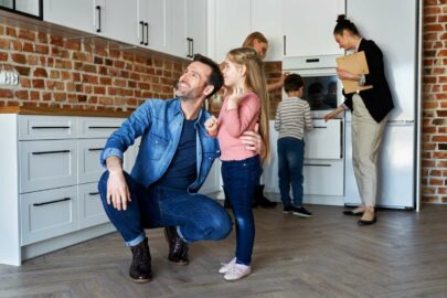 a father crouches down to show his daughter something in the kitchen of their future home while the rest of his family explores behind him
