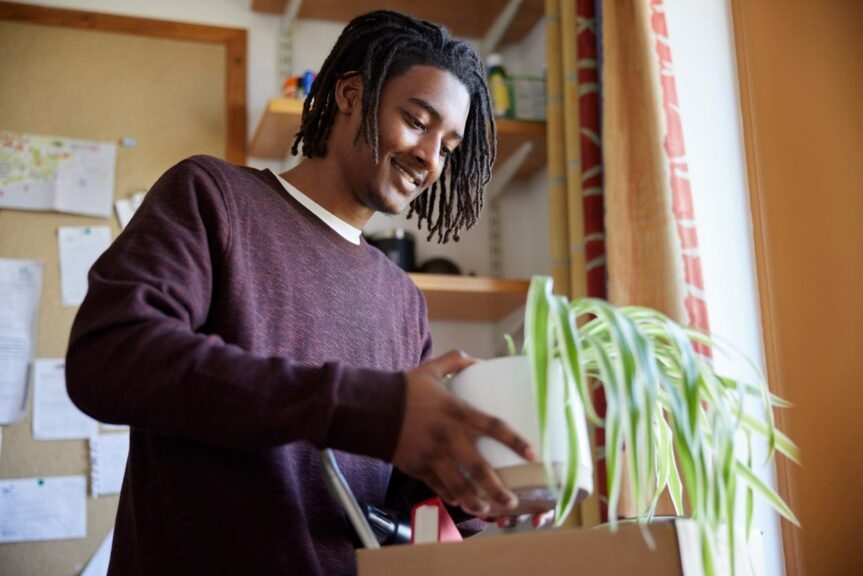 a young man removes a plant from a cardboard box in his dorm room