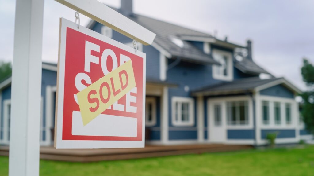 Close up of a For Sale sign in front of a big house. 