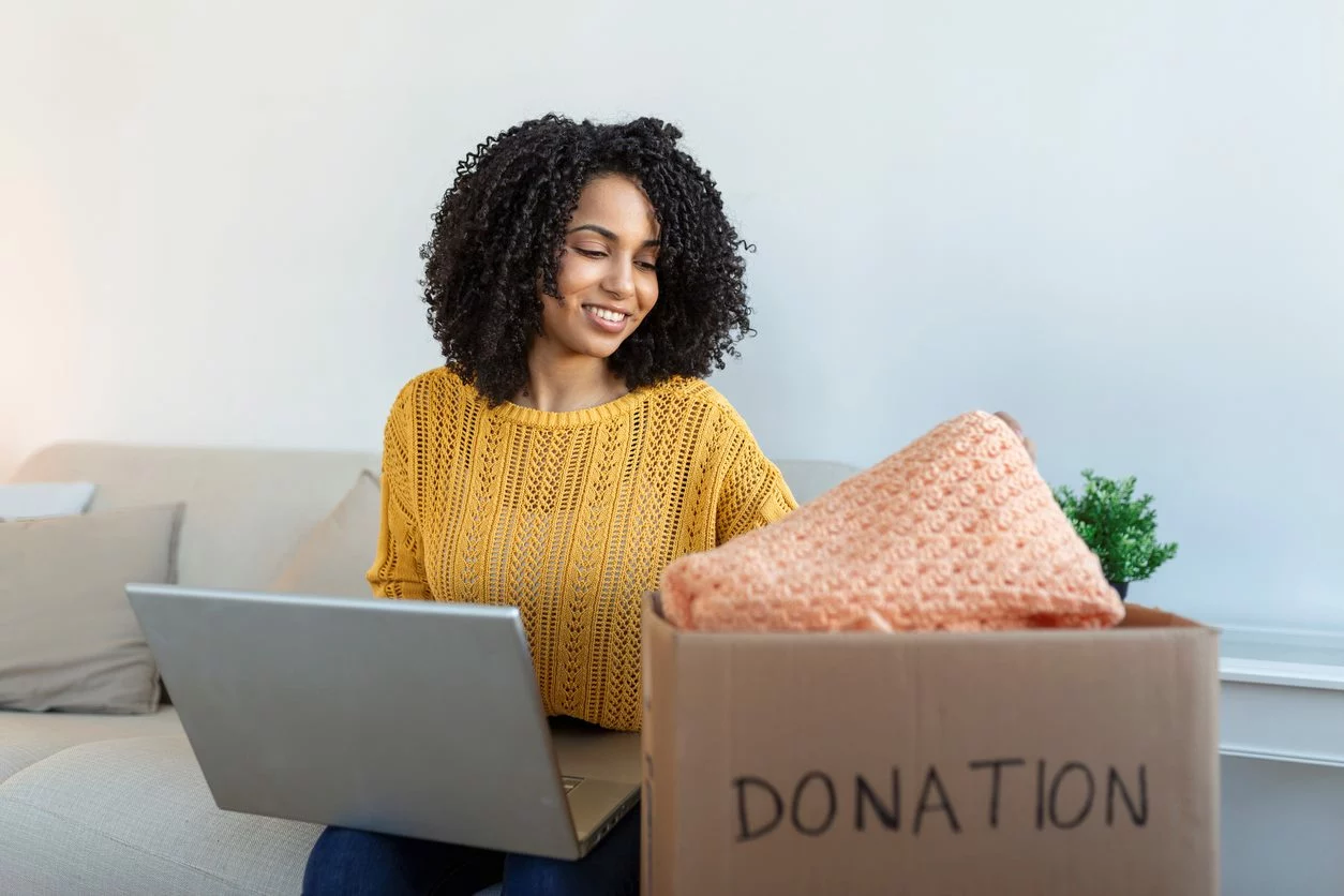 Woman going through a box of donations with a laptop on her lap.