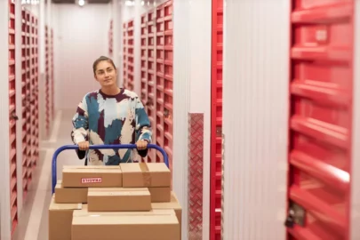 A smiling woman is pushing a large cart filled with cardboard boxes along the corridor of a self storage facility.