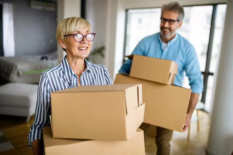 An older man and woman move into a new home, smiling and carrying boxes.