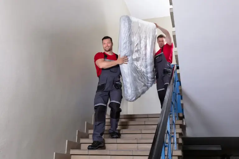 Two professional movers in overalls carry a mattress down a flight of stairs.