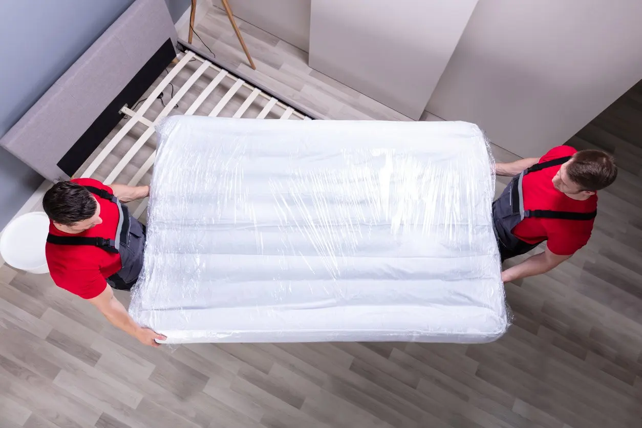 Two movers in red shirts carry a mattress wrapped in plastic out of a room. 