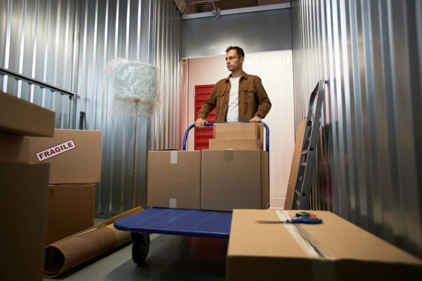 A man in a brown shirt moves boxes inside a 5x10 storage unit with a blue handcart. 